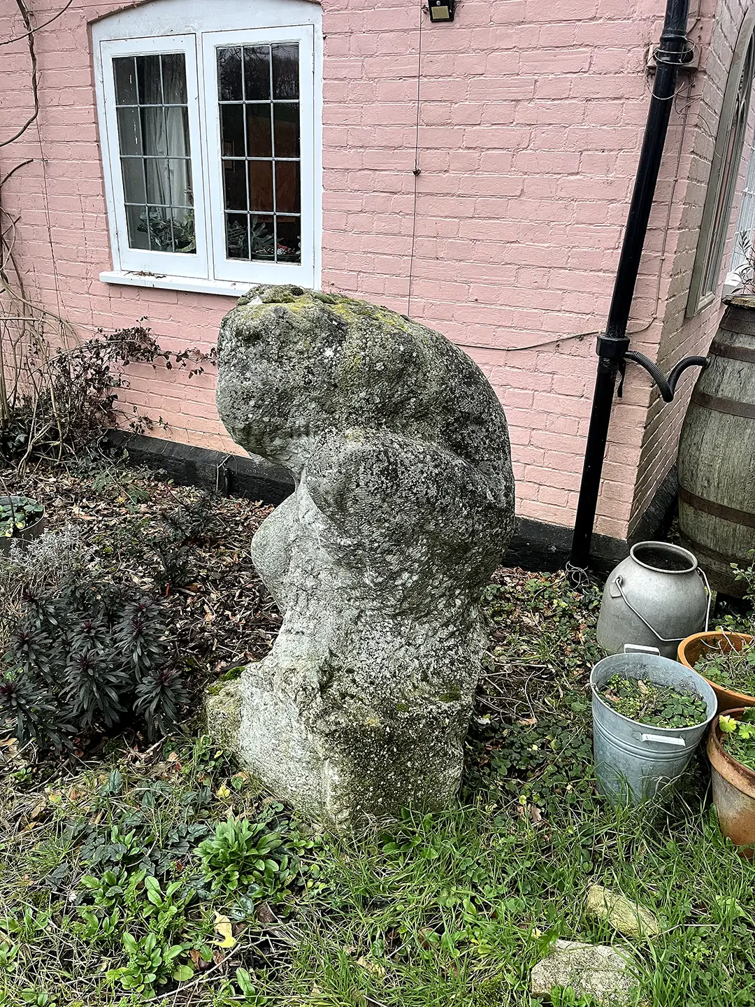 Crouching figure sitting on grass, in front of a pink-painted brick wall.