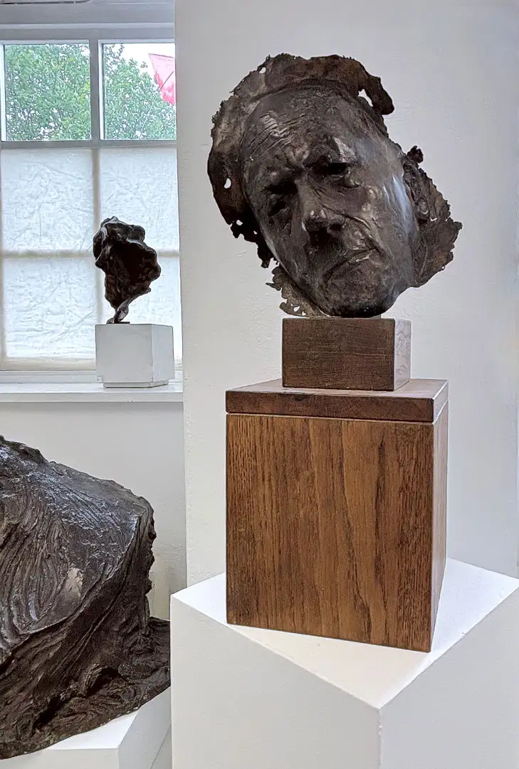 A portraitive sculpture of Alan Rawsthornecast in resin and set onto a wooden base, sitting on a white plinth within a gallery space